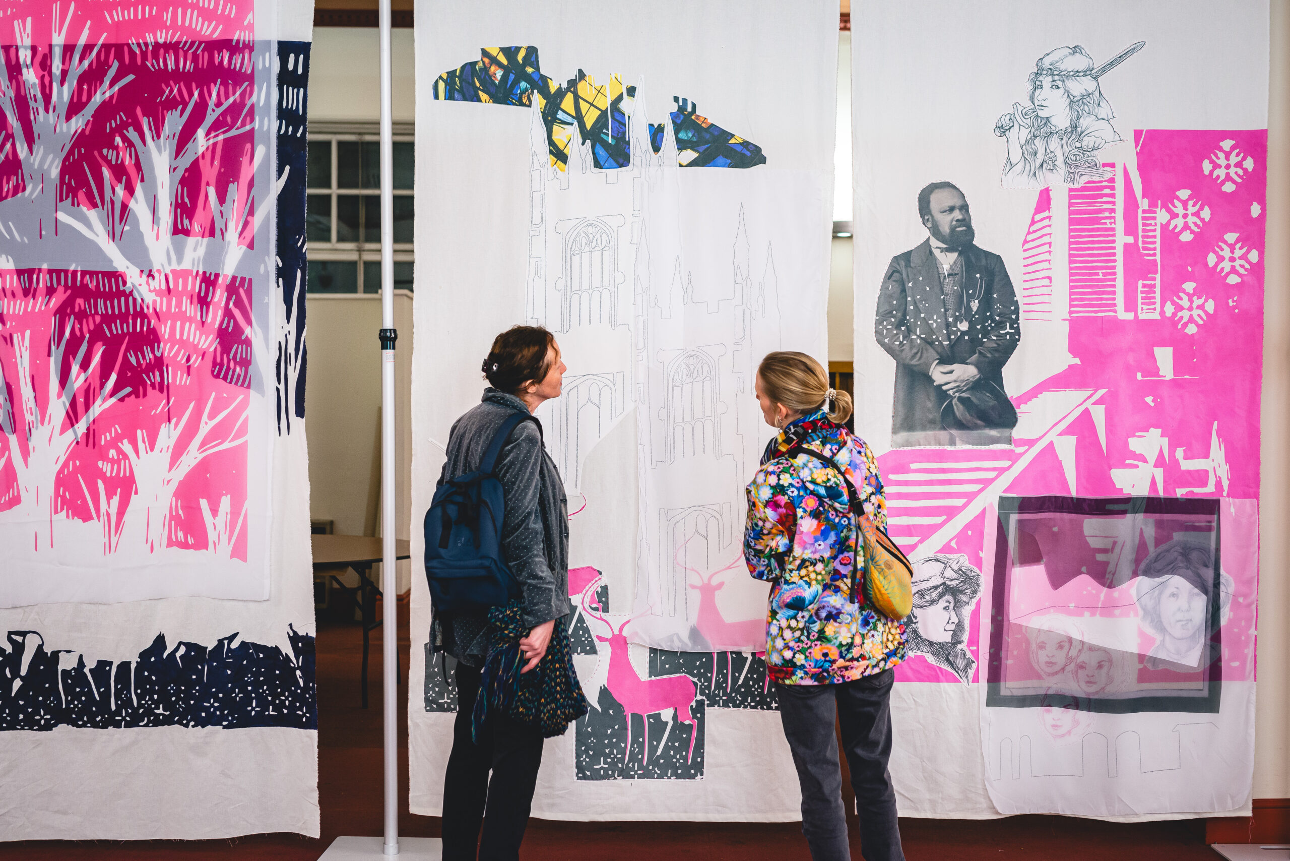 Two people stand in front of a hanging banner which is illustrated with pink and blue, in an exhibition style space